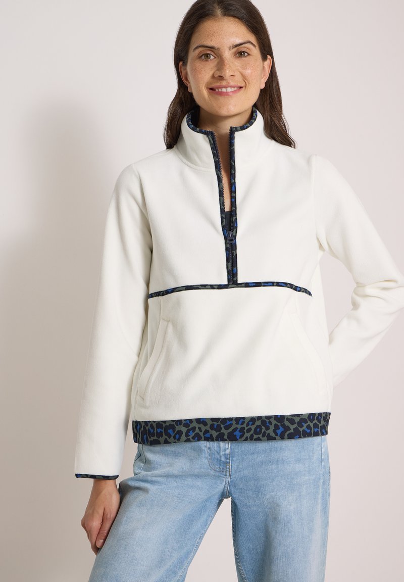 Femme souriante portant un pull blanc avec une bordure imprimé léopard noir et bleu et un jean bleu clair sur un fond clair uni.