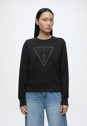 Femme aux cheveux noirs et raides porte un sweat-shirt noir Guess et un jean en denim bleu clair, debout devant un fond blanc uni.