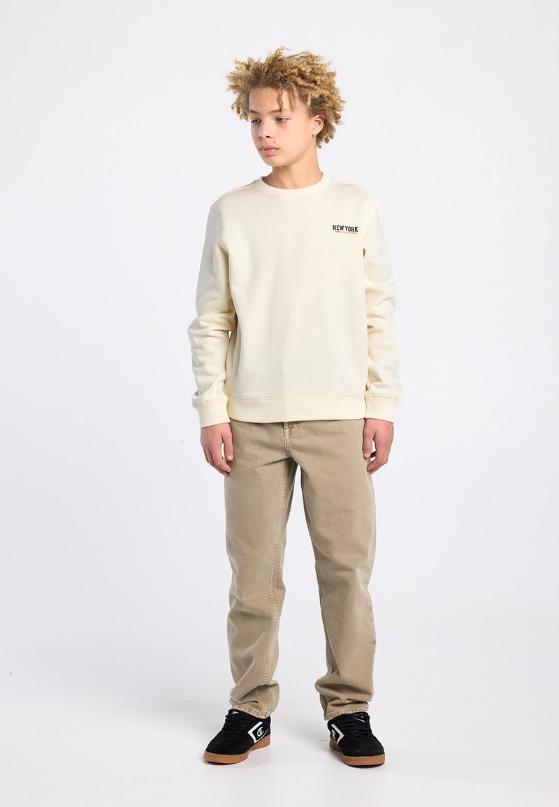 Sweat-shirt crème avec texte « NEW YORK », pantalon khaki coupe droite, et baskets noires. Texture lisse et décontractée.