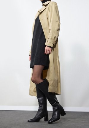 Personne portant un trench-coat beige, une robe-pull col roulé noire, des collants transparents et des bottes à talons hautes noires jusqu'aux genoux sur un fond uni.
