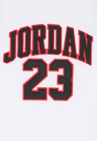 Λευκή φανέλα με το "JORDAN" σε έντονα μαύρα γράμματα με κόκκινο περίγραμμα πάνω από τον αριθμό "23", επίσης μαύρος με κόκκινο περίγραμμα, με υφή από δίχτυ.