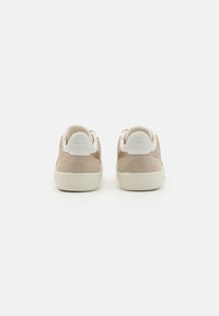 Sneaker in suede beige con dettagli bianchi, punta arrotondata e suola in gomma piatta. La parte posteriore presenta un tallone a contrasto e un branding minimal.