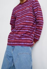 Mörkröd och lila randig långärmad skjorta med rund halsringning och en liten logotyp. Bärs med ljusblå baggy jeans. Texturerad tyg.