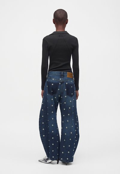 Personne portant un haut noir à manches longues et un jean bleu à jambes larges avec des œillets métalliques, vue de dos sur un fond uni.