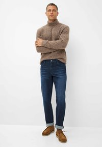 Maglione a collo alto texture marrone, jeans blu scuro e scarpe in camoscio marrone chiaro con risvolti, in piedi su uno sfondo bianco semplice.