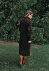 Brun, lang uldfrakke med ribstrikkede ærmer, båret over beige strømpebukser og brune ankelstøvler. Grønne blade i baggrunden.