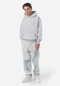 Lichtgrijze hoodie met een buidelzak aan de voorkant, gecombineerd met bijpassende joggers met blauwe golfaccenten en elastische manchetten, gedragen met witte sneakers.