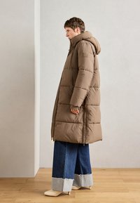 Manteau en duvet marron avec capuche, fermeture éclair et design matelassé. Porté avec un jean à pattes larges présentant un ourlet effiloché et des bottines beige.
