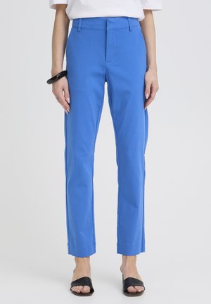 Femme portant un pantalon droit bleu vif, un haut blanc, un bracelet noir et des talons ouverts noirs, debout sur un fond gris clair uni.