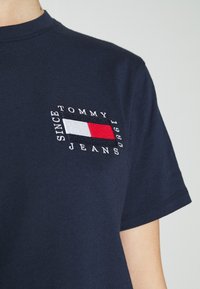 Laivastonsininen puuvillainen t-paita, jossa on kirjailtu logo, jossa on punaisia, valkoisia ja sinisiä elementtejä ja teksti "TOMMY JEANS SINCE 1985." Lyhyet hihat.