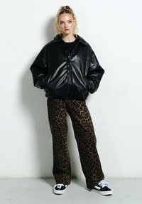 Czarna kurtka z sztucznej skóry w oversize'owym kroju z zapięciem na zamek, zestawiona z brązowymi spodniami w panterkę o szerokich nogawkach oraz czarnymi sneakersami z białą podeszwą.