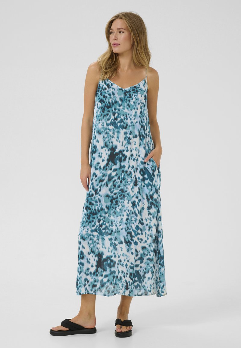 Robe maxi bleue et blanche avec un motif à pois, fabriquée en tissu léger ; dotée de fines bretelles et de poches latérales. Portée avec des sandales noires.