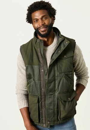 Man with beard wearing green utility vest over beige sweater, smiling with one hand in pocket against plain light background.