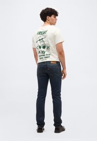 Νέος άνδρας που φοράει λευκό t-shirt με πράσινα γράμματα και σχέδια, σκούρα μπλε τζιν και μαύρα παπούτσια, γυρισμένος από πίσω με το κεφάλι στραμμένο προς τα δεξιά.