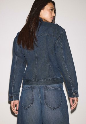 Femme aux longs cheveux foncés portant une veste en jean bleu marine et un jean assorti, tournée de dos et regardant vers sa droite.