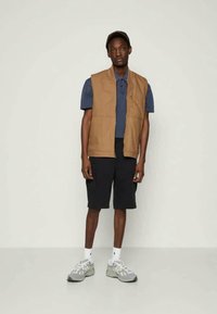 Tan spalvos liemenė ant mėlynos polo marškinėlių, derinama su juodomis šortais ir pilkais sportbačiais. Liemenė turi užtrauktuką ir laisvą kirpimą.