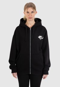 Veste à capuche zippée noire avec une grande poche avant, cordon de serrage à la capuche et motif graphique blanc sur le côté gauche de la poitrine. Texture douce, coupe oversize.
