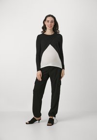Top nero a maniche lunghe sovrapposto a uno strato bianco, abbinato a pantaloni cargo neri. La modella indossa mule nere con tacco, sorridendo.