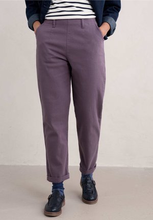 Persona con pantalones morados remangados, zapatos azul marino con hebillas, calcetines azules, camiseta a rayas y chaqueta oscura con las manos en los bolsillos.