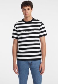 Černobíle pruhované tričko s kulatým výstřihem; uprostřed je logo "GUESS JEANS"; kombinováno s džínovými kalhotami.