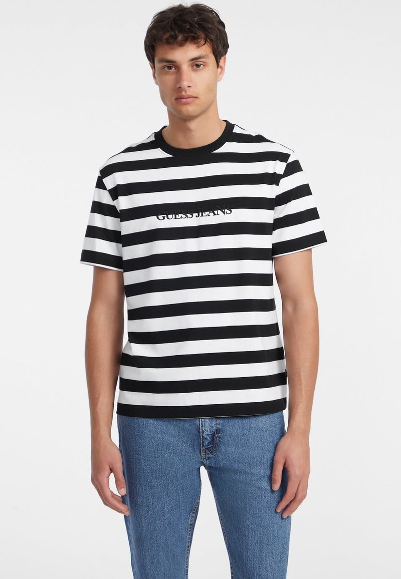 Černobíle pruhované tričko s kulatým výstřihem; uprostřed je logo "GUESS JEANS"; kombinováno s džínovými kalhotami.