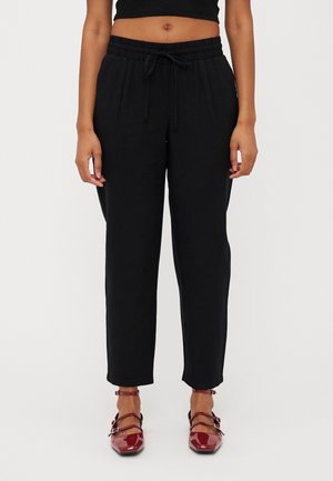 Vrouw staand, draagt zwarte broek met trekkoord en glimmende rode schoenen met gesp, kort zwart topje, tegen een effen lichte achtergrond.