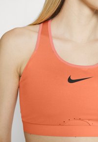 Sutiã desportivo laranja com pormenores em rosa, apresentando um logo da Nike em preto. Feito de um tecido suave, inclui detalhes em malha respirável na parte inferior.