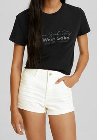 Camiseta de algodón negra con mangas cortas, que presenta una impresión gráfica que dice "New York City West Soho" y unos pantalones cortos de mezclilla blanca con dobladillos enrollados.