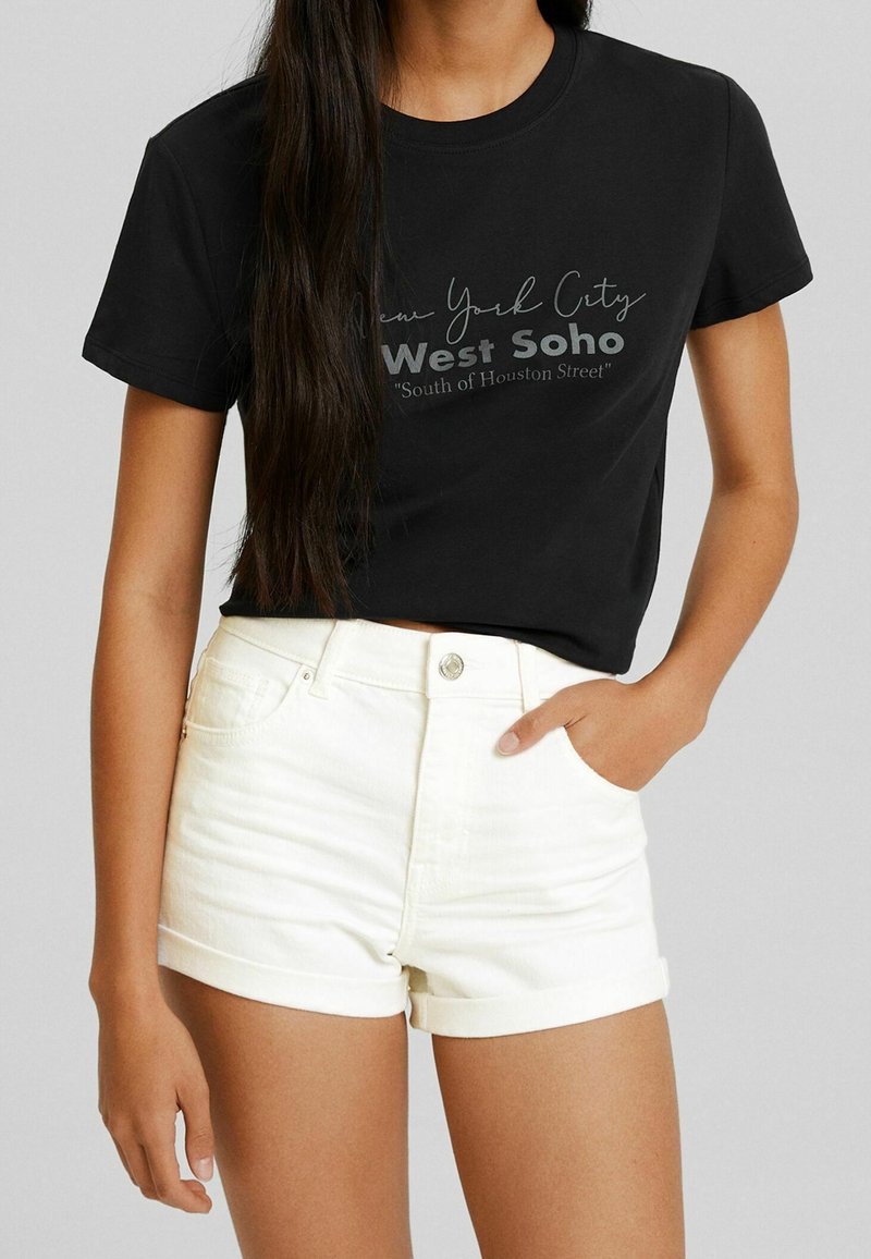 Camiseta de algodón negra con mangas cortas, que presenta una impresión gráfica que dice "New York City West Soho" y unos pantalones cortos de mezclilla blanca con dobladillos enrollados.