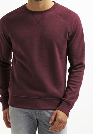 Hombre vistiendo una sudadera de cuello redondo lisa color granate con puños y dobladillo acanalados, combinada con jeans gris claro, de pie contra un fondo blanco.