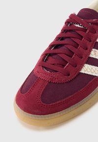 Αθλητικό παπούτσι από μπορντό σουέντ με κρεμ ρίγες, με στρογγυλή μύτη και υφασμένο πέλμα από λάστιχο γκαμ. Σχέδιο με κορδόνια και ενισχυμένες ραφές.