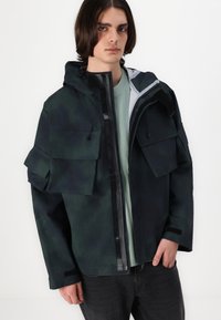 Hooded jacket in dark green and black with multiple large pockets. Features a zip-front closure and elastic drawcords for adjustments.