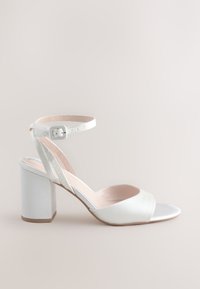 Hvid satin-hæl med åben tå, ankelrem og blokhæl. Har en glat tekstur og en sølvaccent på tåen.
