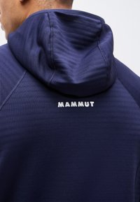 Άνδρας που φοράει μπλε ναυτικό φούτερ με κουκούλα, με το λογότυπο "MAMMUT" στην πάνω πλάτη, εμφανίζοντας λεπτομερή ραφές και υφή υφάσματος.