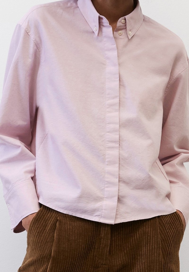 Personne portant une chemise rose pâle boutonnée rentrée dans un pantalon en velours côtelé marron taille haute, les mains dans les poches.