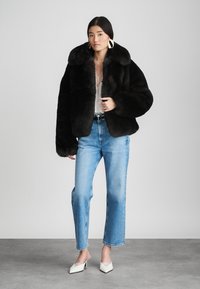 Μαύρο μπουφάν από ψεύτικη γούνα με φαρδύ γιακά, φορεμένο πάνω από ένα ανοιχτόχρωμο τοπ και μπλε τζιν καλή γραμμή. Το μοντέλο στέκεται σε γκρίζο πάτωμα, φορώντας λευκά τακούνια.