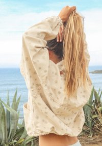 Veste beige clair avec un motif floral, des poignets élastiques et une coupe ample ; possède une poche avant et un design décontracté.