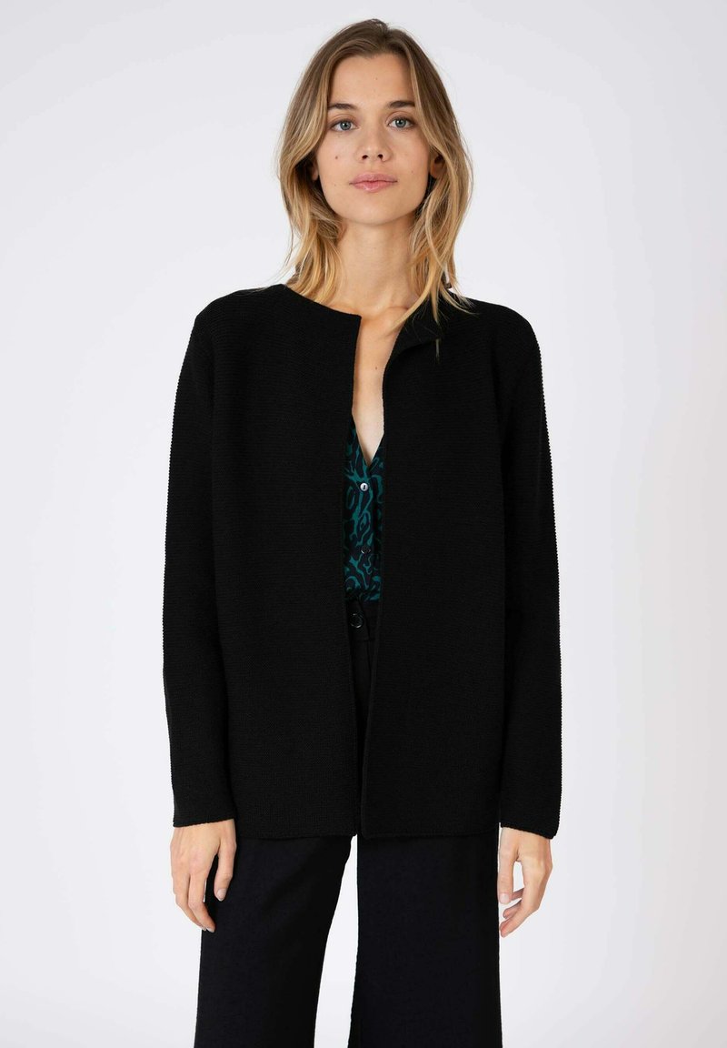 Gilet noir en tricot avec un col rond et une ouverture sur le devant. Le tissu a une finition texturée et arrive jusqu'à la taille.