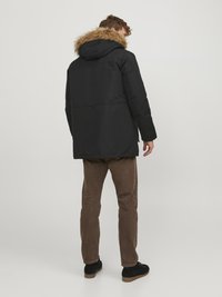 Juodas parka tipo švarkas su dirbtinio kailio gaubtu, sukurtas su tiesia siluetu. Modelis dėvi rudas kordūrozo kelnes ir juodus batus.