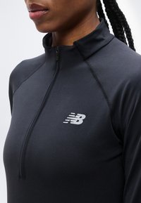 Κοντινό πλάνο ενός ατόμου που φοράει μαύρο αθλητικό τοπ με φερμουάρ και λευκό λογότυπο New Balance στο στήθος και πλεγμένα μαλλιά.