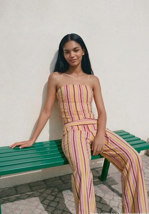 Młoda kobieta o długich ciemnych włosach, ubrana w żółty, różowy i biały prążkowany top bez ramiączek oraz spodnie, siedzi na zielonej ławce na tle białej ściany.