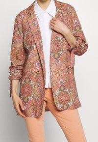 Blazer à motifs dans des tons vibrants de rose, orange et or. Tissu léger avec un motif floral, bouton unique et manches retroussées.