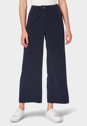 Femme portant un pantalon large court bleu marine avec plis devant, bouton et fermeture éclair, associé à des baskets blanches et un haut blanc.