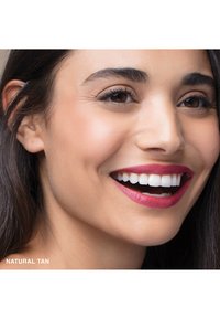 Viso sorridente con pelle di media abbronzatura, capelli scuri e denti bianchi brillanti. Le labbra sono lucide, ricoperte di una tonalità di rosa intenso. Le sopracciglia sono ben definite.