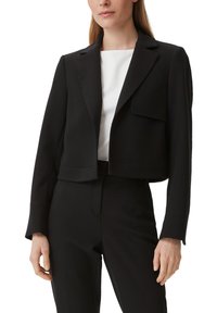 Zwarte cropped jas met een notch rever, klepzakken en lange mouwen. Gemaakt van een gladde, gestructureerde stof. Draagt over een witte top.
