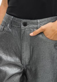 Pantalones metálico gris con un bolsillo frontal, cierre de botón y textura suave. El diseño presenta una silueta elegante y moderna.