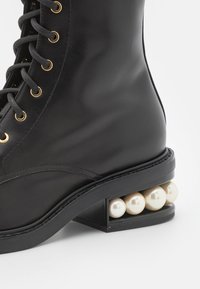 Botín negro de cuero con ojales dorados, cordones grises y un tacón cuadrado que presenta acentos de perlas blancas. Textura suave y diseño redondeado.