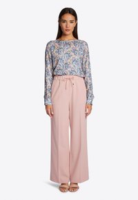 Langärmlige Bluse mit floralem Druck in Pink, Blau und Grün; kombiniert mit weit geschnittenen rosa Hosen und neutralfarbenen Sandalen.