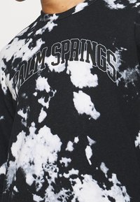 La camiseta de manga larga en blanco y negro con efecto tie-dye presenta el texto "Palm Springs" en letras negras curvas en el pecho. Tela de algodón, diseño de cuello redondo.