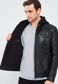 Chaqueta de cuero negra con un diseño acolchonado, bolsillos frontales con cremallera y una capucha negra desmontable. Llevada sobre una camiseta gris clara.